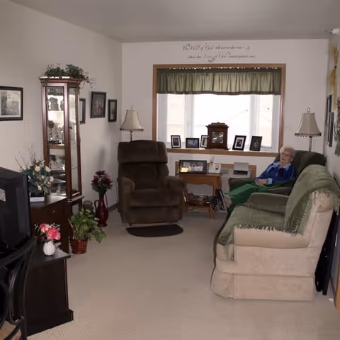 A cozy living room with beige carpet, a beige sofa with a green throw, a brown recliner chair, and a wooden display cabinet with glass doors. There are two table lamps on either side of the window, which has a green valance. A small wooden table under the window holds framed photos and a clock. An elderly person is sitting on the sofa. The walls are decorated with framed pictures and a wall decal above the window.