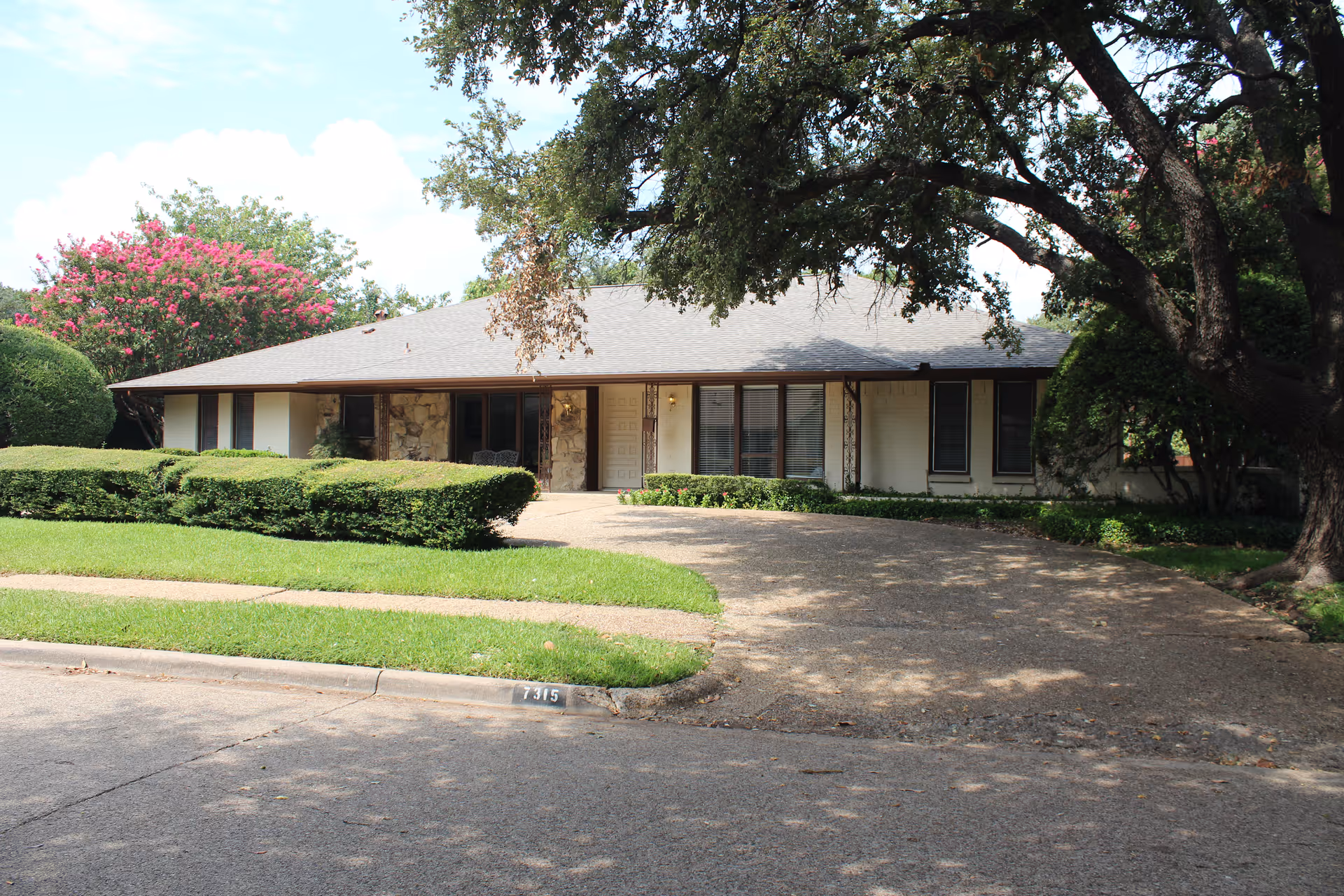 Single-story residential building with a sloped roof, stone and light-colored exterior walls, surrounded by well-maintained green bushes and trees. A curved driveway leads to the front entrance, and the house number 7315 is visible on the curb.