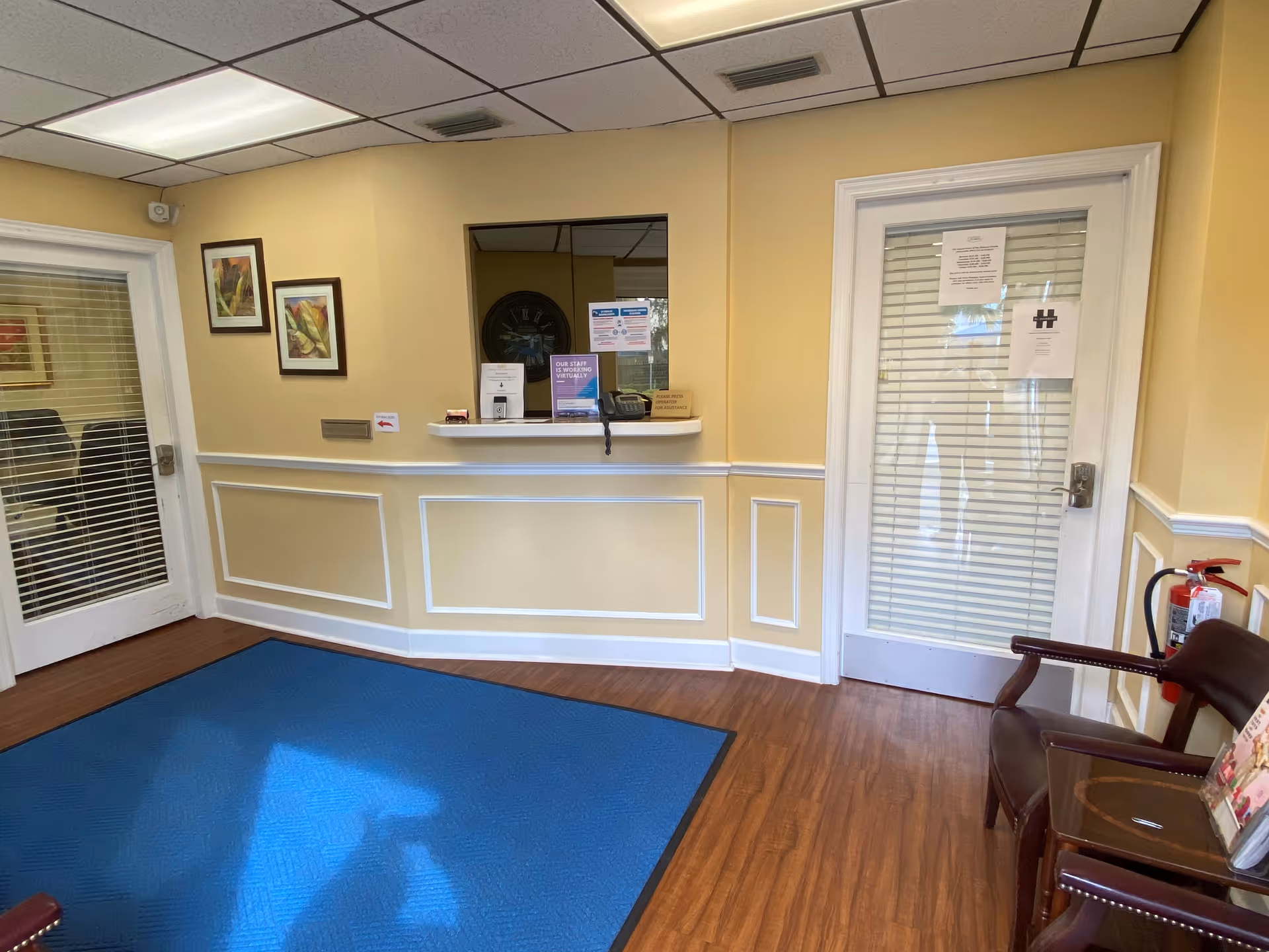 Reception desk with doorway window, chairs, and blue mat in a yellow-walled facility lobby.
