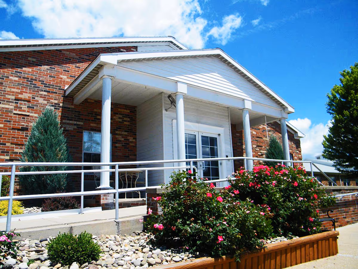 Entrance of a brick building with white columns and a covered porch. There is a wheelchair accessible ramp with metal railings leading to double glass doors. The area is landscaped with bushes, flowers, and small trees under a blue sky with some clouds.