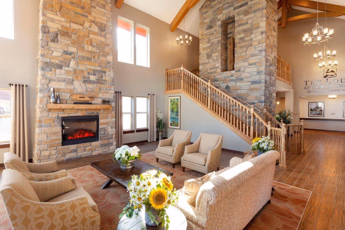 A spacious living room area with high ceilings and large windows letting in natural light. The room features a tall stone fireplace with a glowing fire, beige upholstered chairs and sofas arranged around a wooden coffee table with flower arrangements. A wooden staircase with a railing leads to an upper level. The walls are decorated with framed artwork and chandeliers hang from the ceiling. The far wall displays a sign that reads 'The Lodge at North Ogden'.