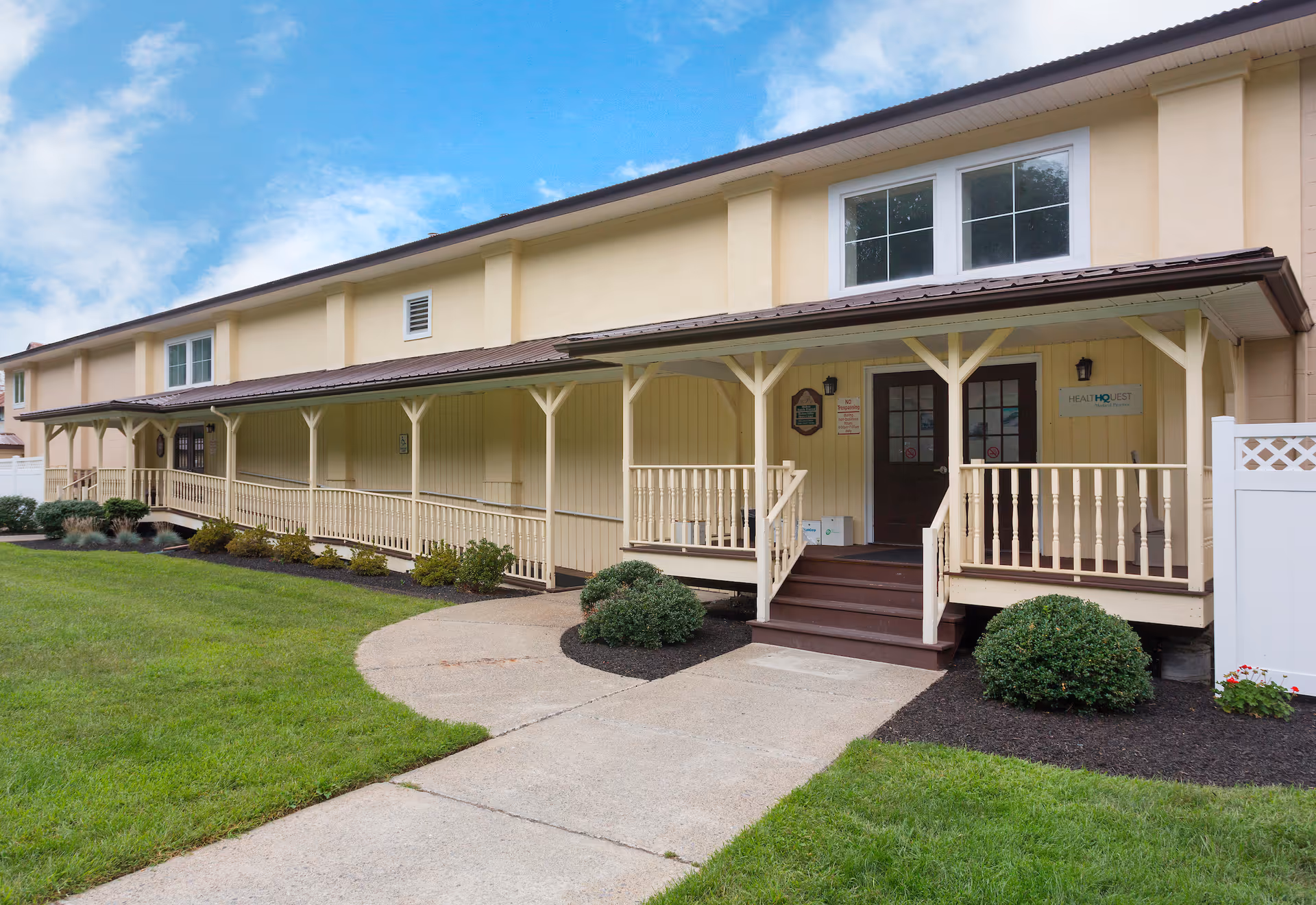 Two-story beige senior living building with a covered front porch, walkway, and manicured lawn.