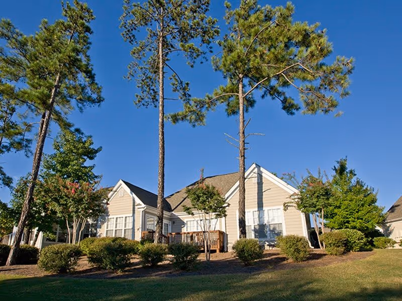 Exterior view of a single-story residential building with beige siding and white trim, surrounded by tall pine trees, bushes, and a well-maintained lawn under a clear blue sky.