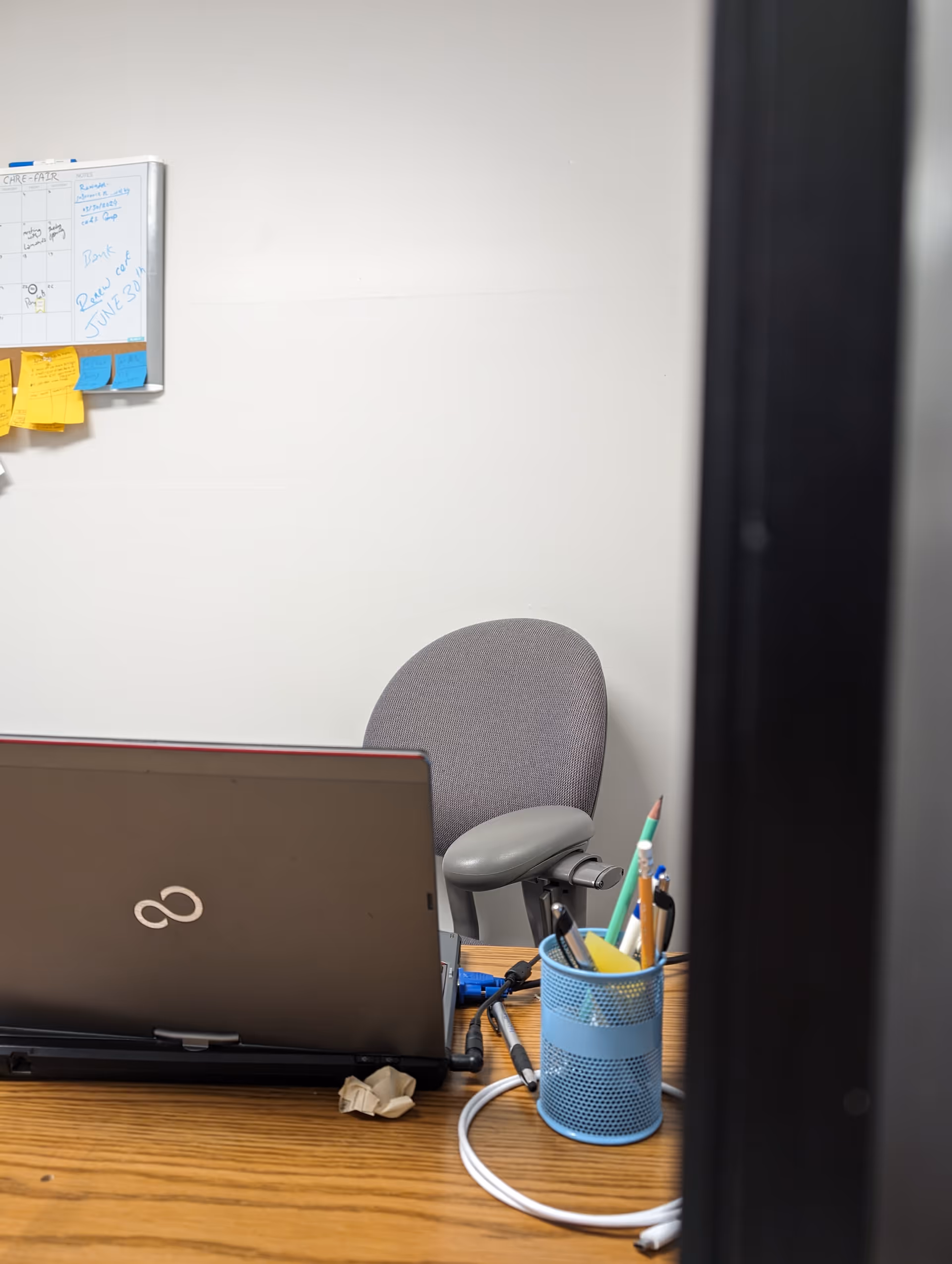 An office desk featuring a laptop, blue pencil cup, and sticky notes beside a whiteboard calendar.