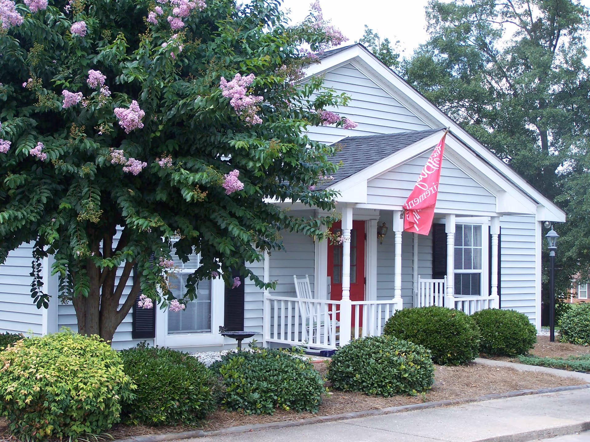 Front of a small light-gray house with a white porch, red door and flag, surrounded by shrubs and a blooming pink-flowered tree.