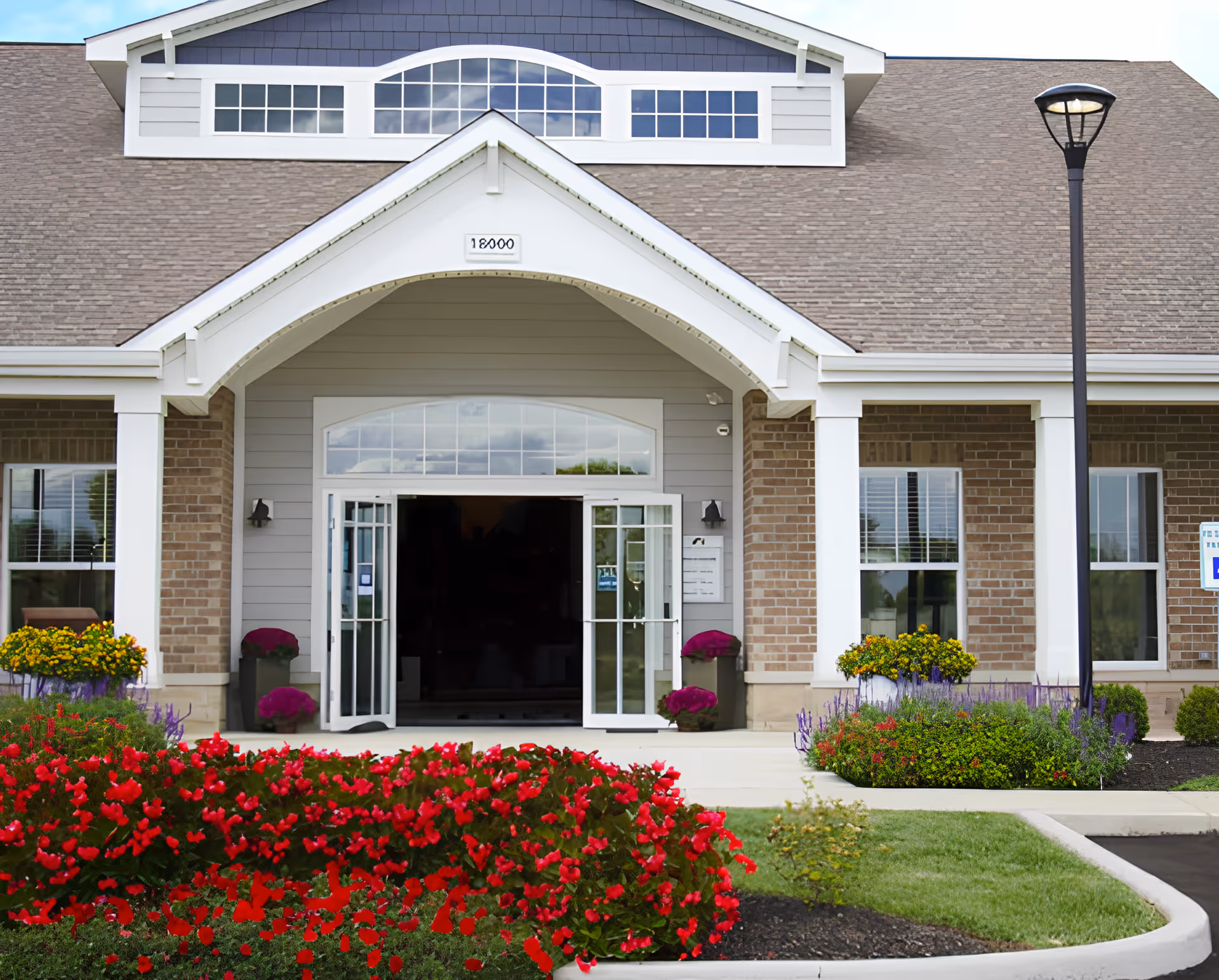 Front exterior view of Promenade Apartments building entrance with a covered porch, glass double doors, brick and siding facade, and landscaped flower beds with red and yellow flowers in front.