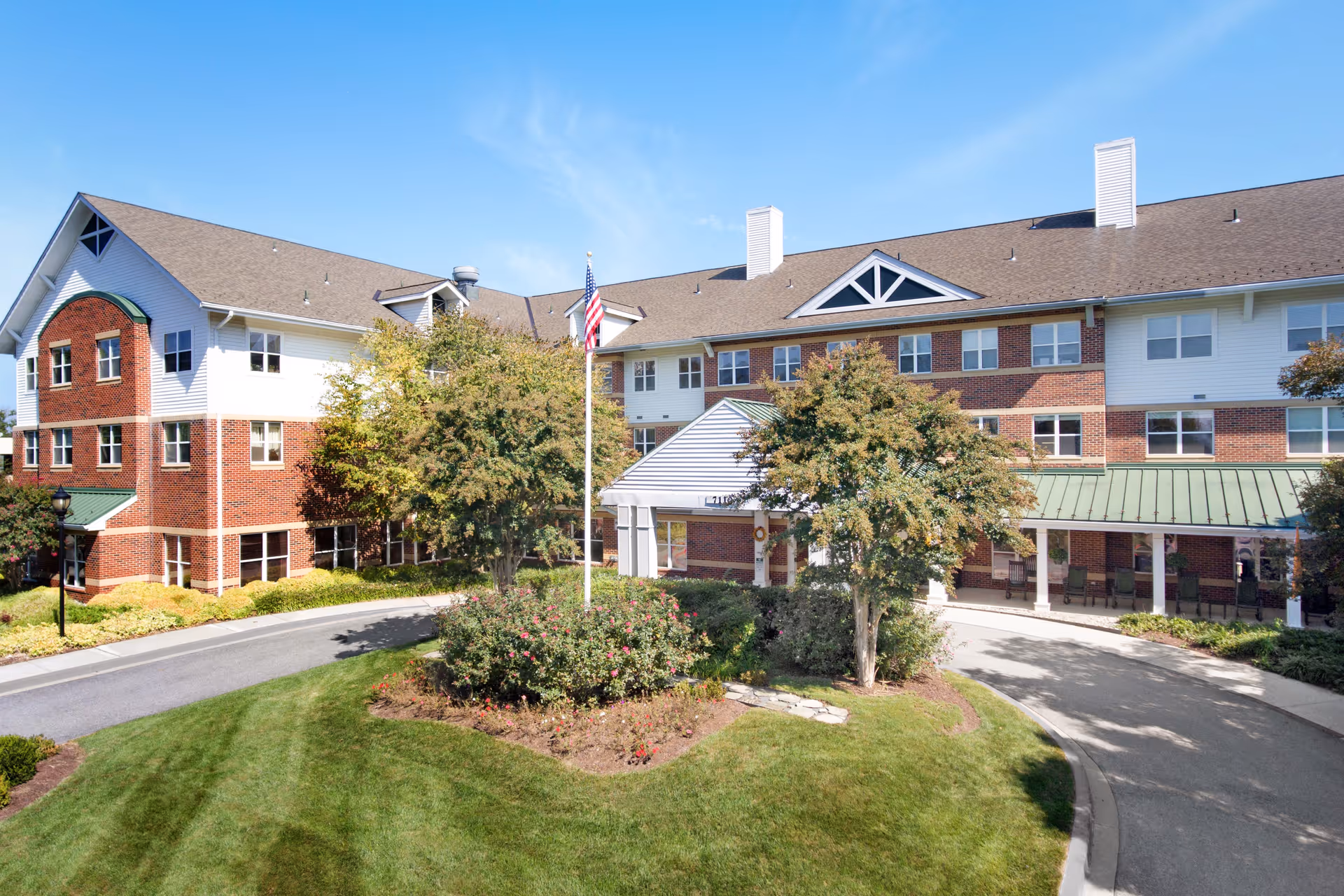 Front exterior of a three-story brick-and-siding senior living building with a circular driveway, flagpole, and landscaped entrance.