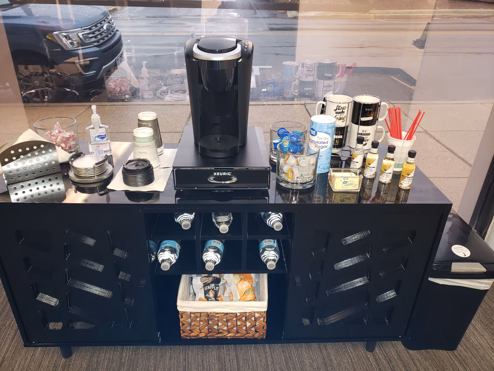Coffee station with Keurig, condiments, cups, and assorted syrups on a dark cabinet.