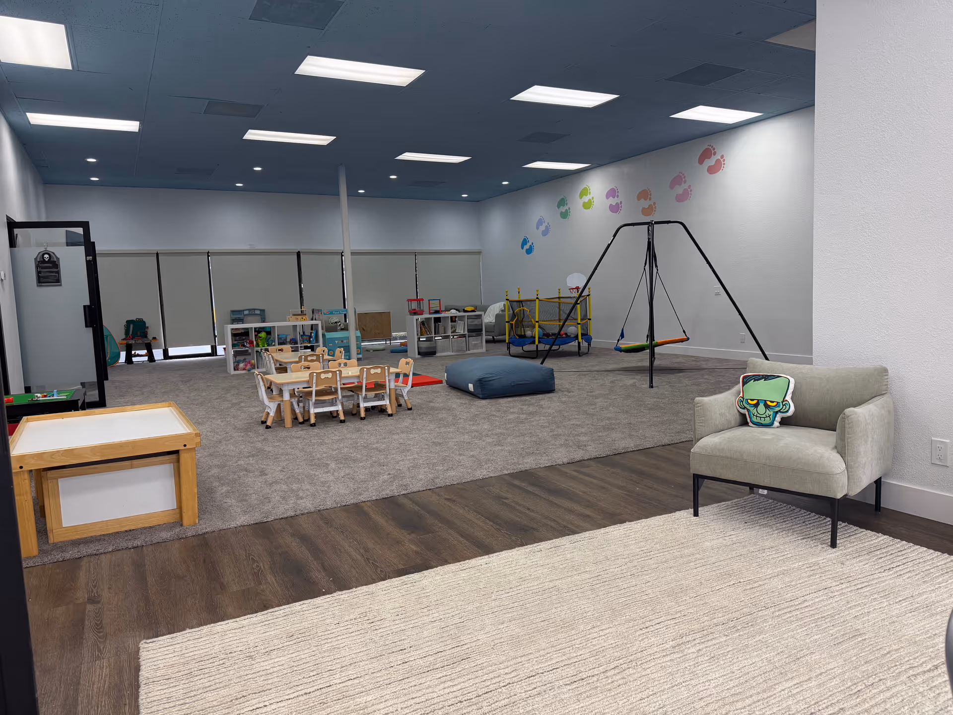 Indoor playroom with small tables, chairs, toys, a swing set, padded mats, and a lounge chair.