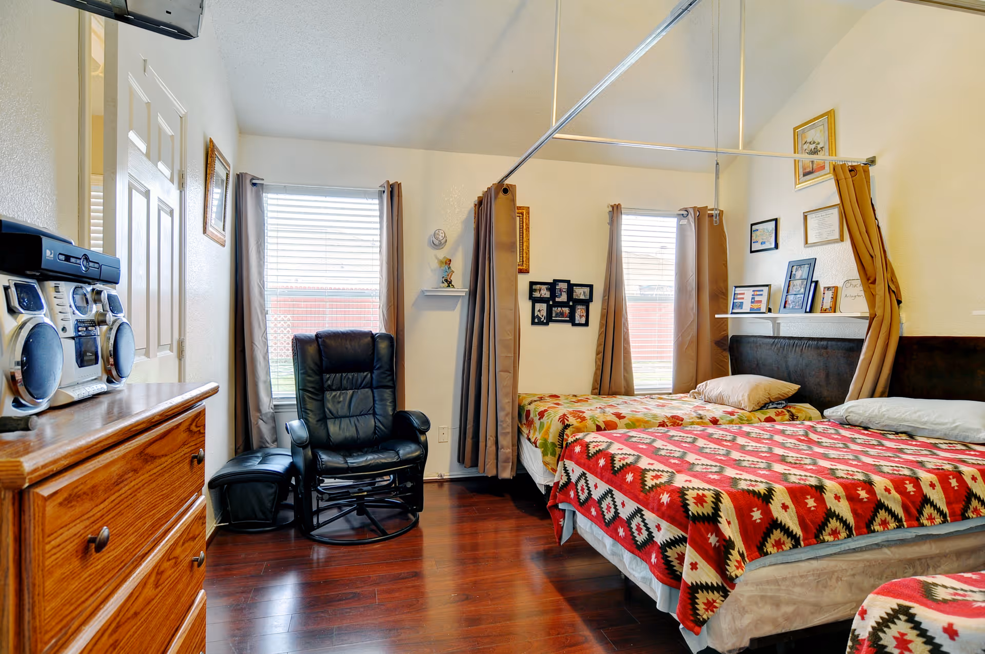 A cozy bedroom with two single beds covered in colorful patterned blankets. The room has wooden flooring, two windows with brown curtains, a black leather recliner chair, a wooden dresser with a stereo system on top, and various framed pictures and certificates on the walls.