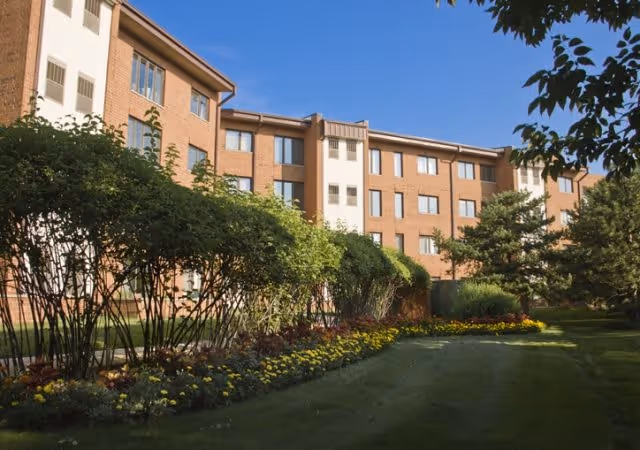 Exterior view of a multi-story brick building with several windows, surrounded by well-maintained landscaping including bushes, trees, and flower beds under a clear sky.