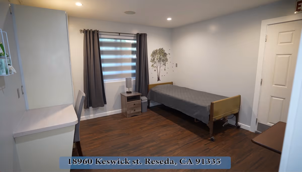 A simple assisted-living bedroom with a single bed, nightstand, desk, and window with gray curtains.