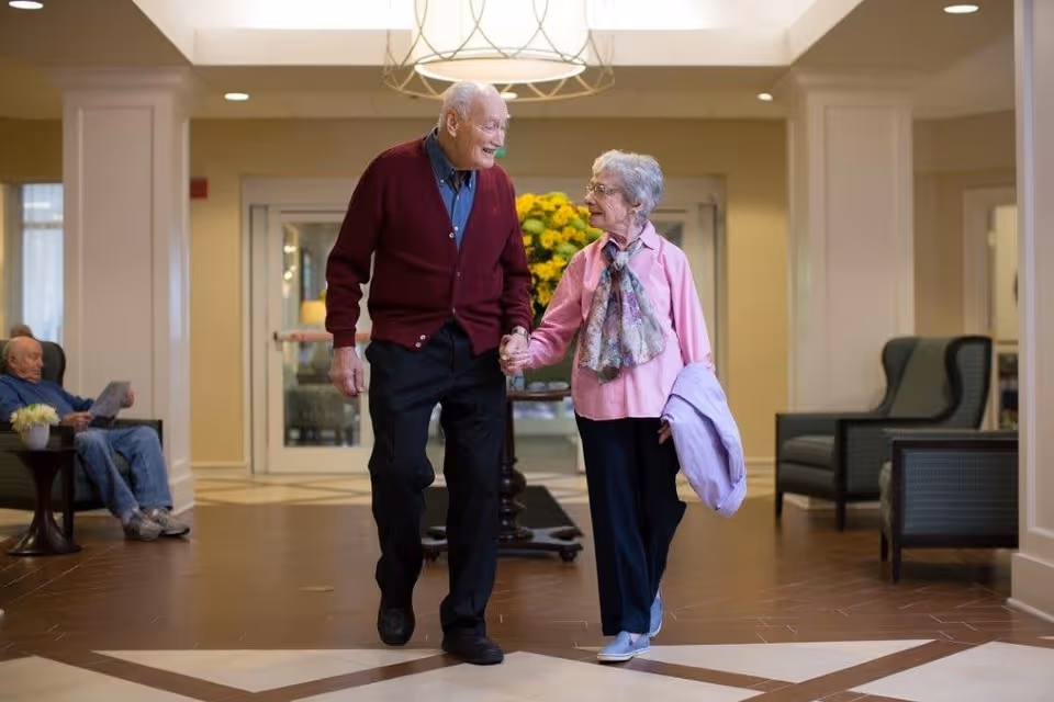 An elderly couple holding hands and walking together in a well-lit hallway of a senior living facility. The man is wearing a maroon cardigan and black pants, while the woman is dressed in a pink blouse with a floral scarf and holding a light purple jacket. In the background, a man is sitting on a chair reading a newspaper, and there are armchairs and a table with flowers along the walls.