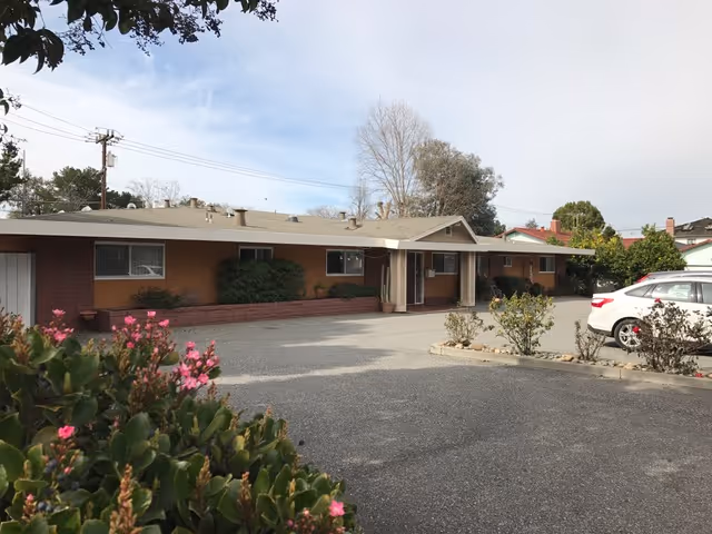 Single-story tan building with a covered front entrance, parking lot, and flowering shrubs in the foreground.