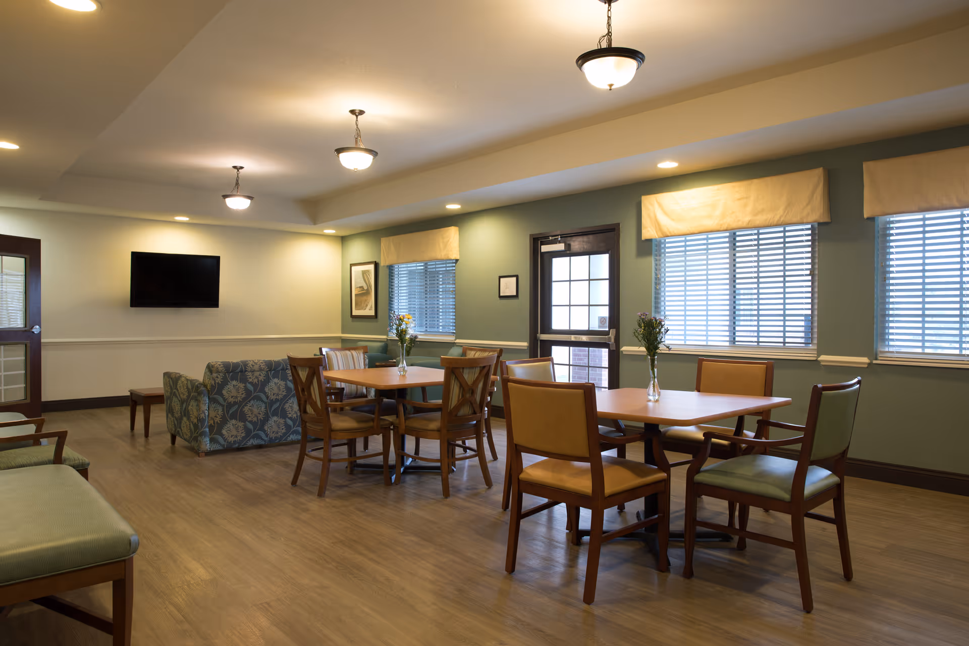 A well-lit senior living dining area with tables, chairs, a blue sofa, and windows.