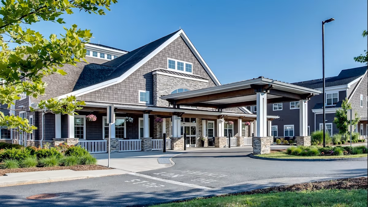 Front exterior view of Artisan at Hudson facility showing a large covered entrance with stone pillars, a driveway, landscaped greenery, and clear blue sky.