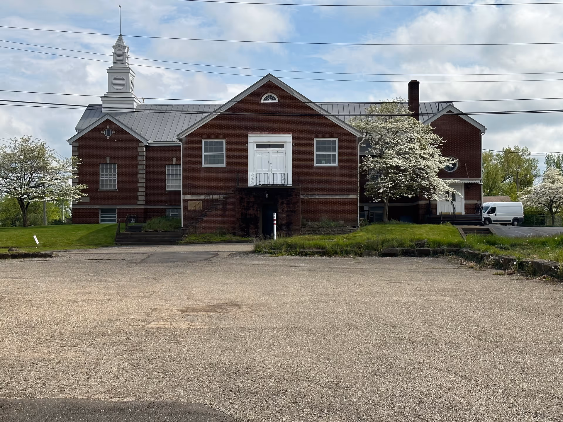 Brick building with a white cupola, blooming trees, and a white van in the parking lot.