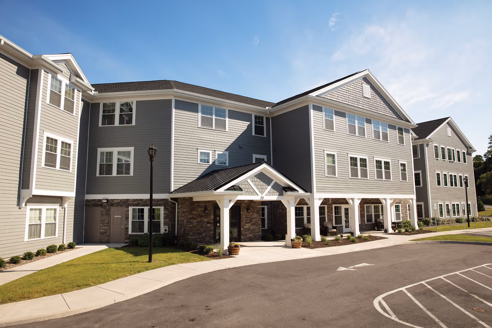 Exterior view of The Woodlands Senior Apartments building with gray siding, white trim, multiple windows, a covered entrance, and a parking area in front under a clear blue sky.