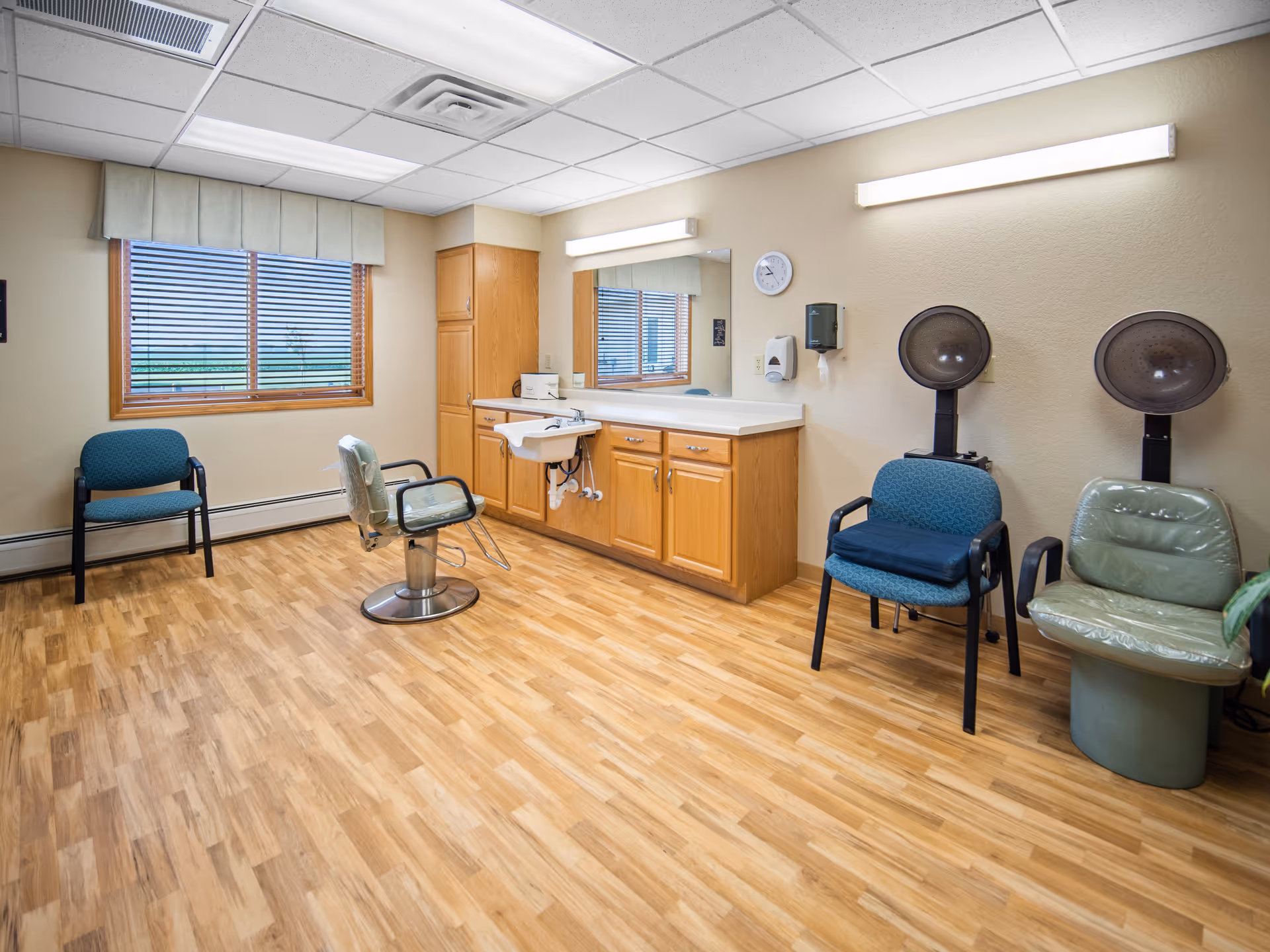 Interior room with wooden flooring and beige walls featuring a large mirror above a countertop with cabinets and a sink. There are two hair dryer chairs on the right side, a salon chair in the center, and two blue chairs near a window with blinds on the left side. The ceiling has fluorescent lighting panels.