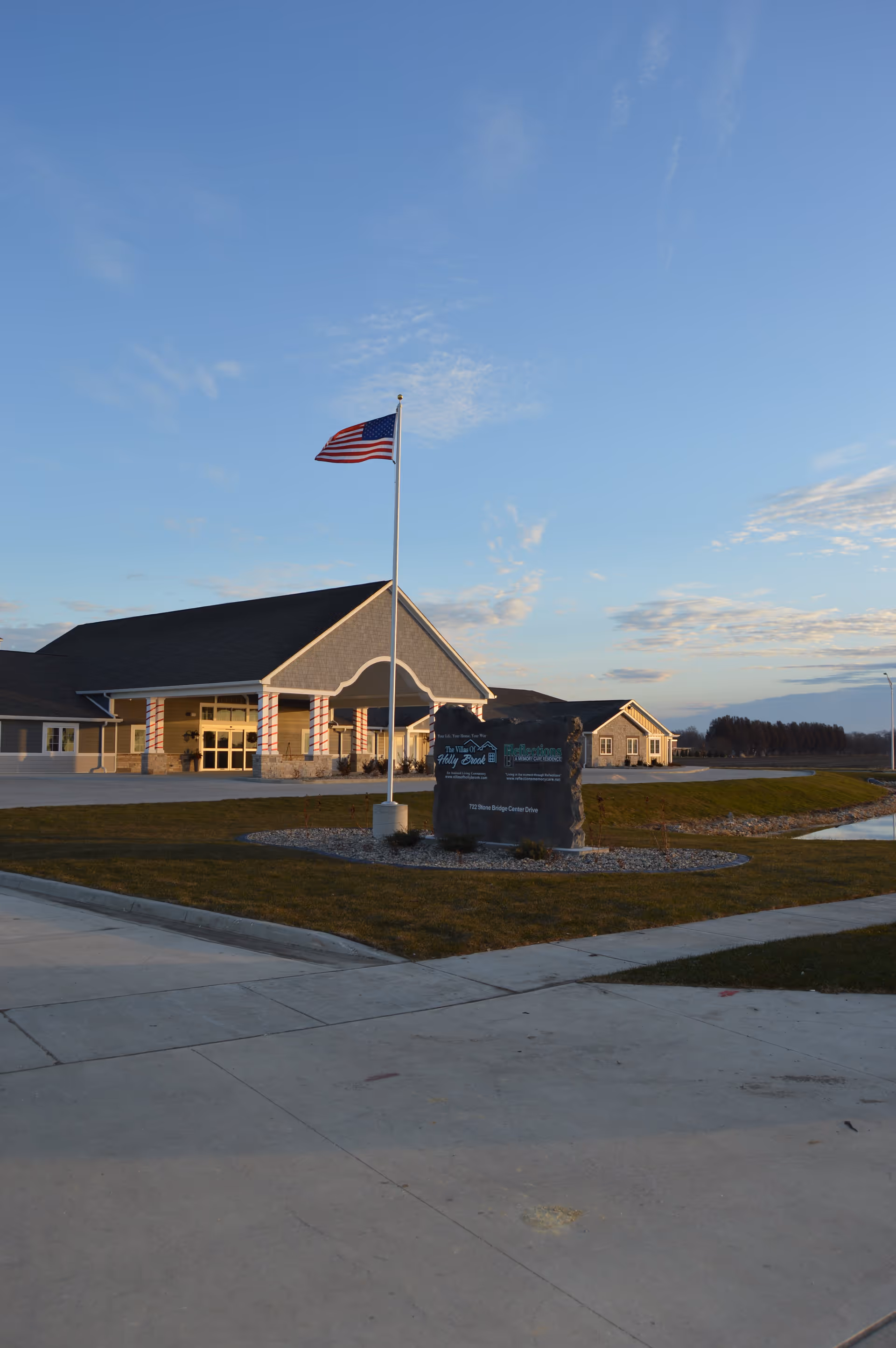 Exterior view of Villas of Holly Brook Assisted Living & Memory Care building with a flagpole flying the American flag in front. The building has a covered entrance with columns wrapped in red and white stripes. A stone sign with the facility's name and address is visible near the entrance, along with a small landscaped area and a pond to the right.
