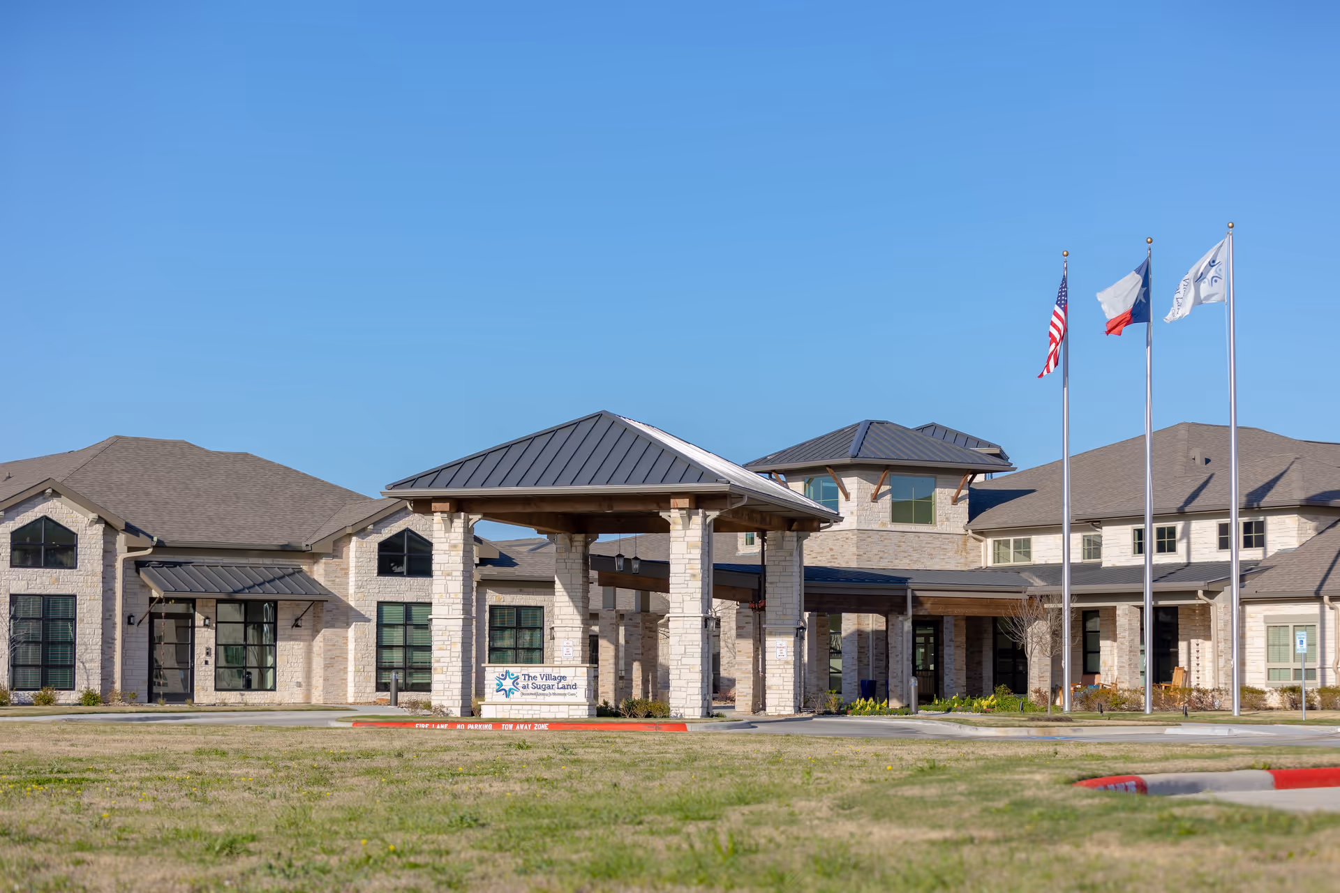 Exterior view of The Village at Sugar Land senior living facility with a covered entrance, stone facade, and three flagpoles displaying the American flag, Texas state flag, and the facility flag under a clear blue sky.