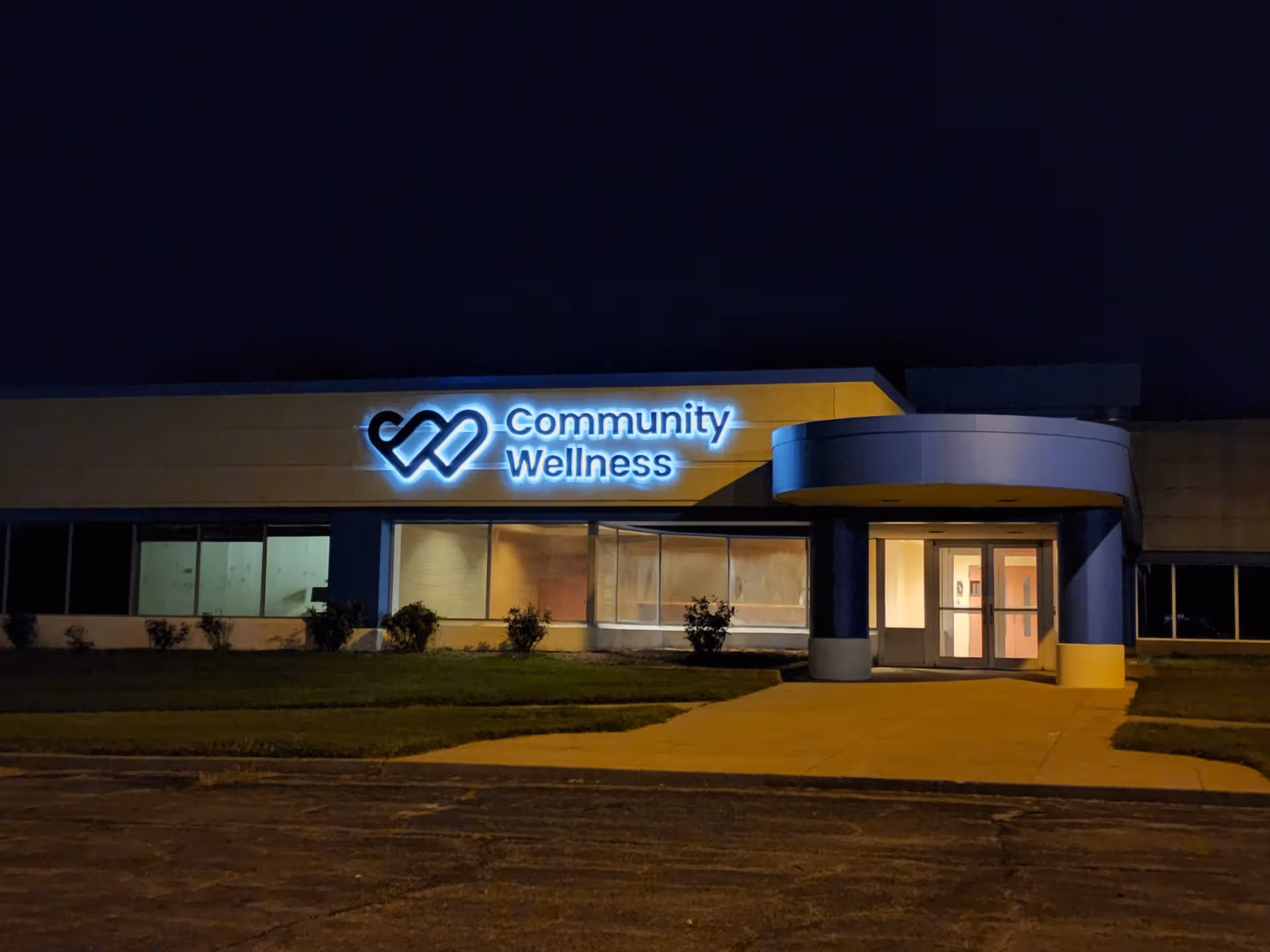 Night exterior of a facility with an illuminated Community Wellness sign at the entrance.