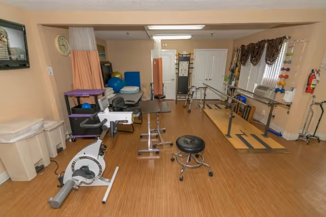 A rehabilitation room with exercise equipment including a stationary bike, parallel bars for walking practice, therapy tables, exercise balls, and various physical therapy tools. The room has wooden flooring, beige walls, and windows with brown curtains. There is a clock on the wall and a television mounted in the corner.