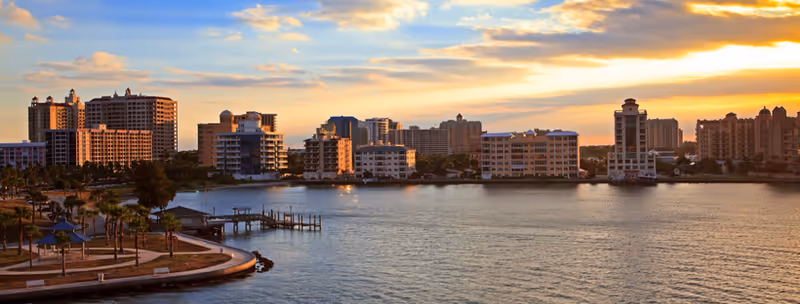 Sunset over a waterfront cityscape with river, palm trees, and a curved promenade.