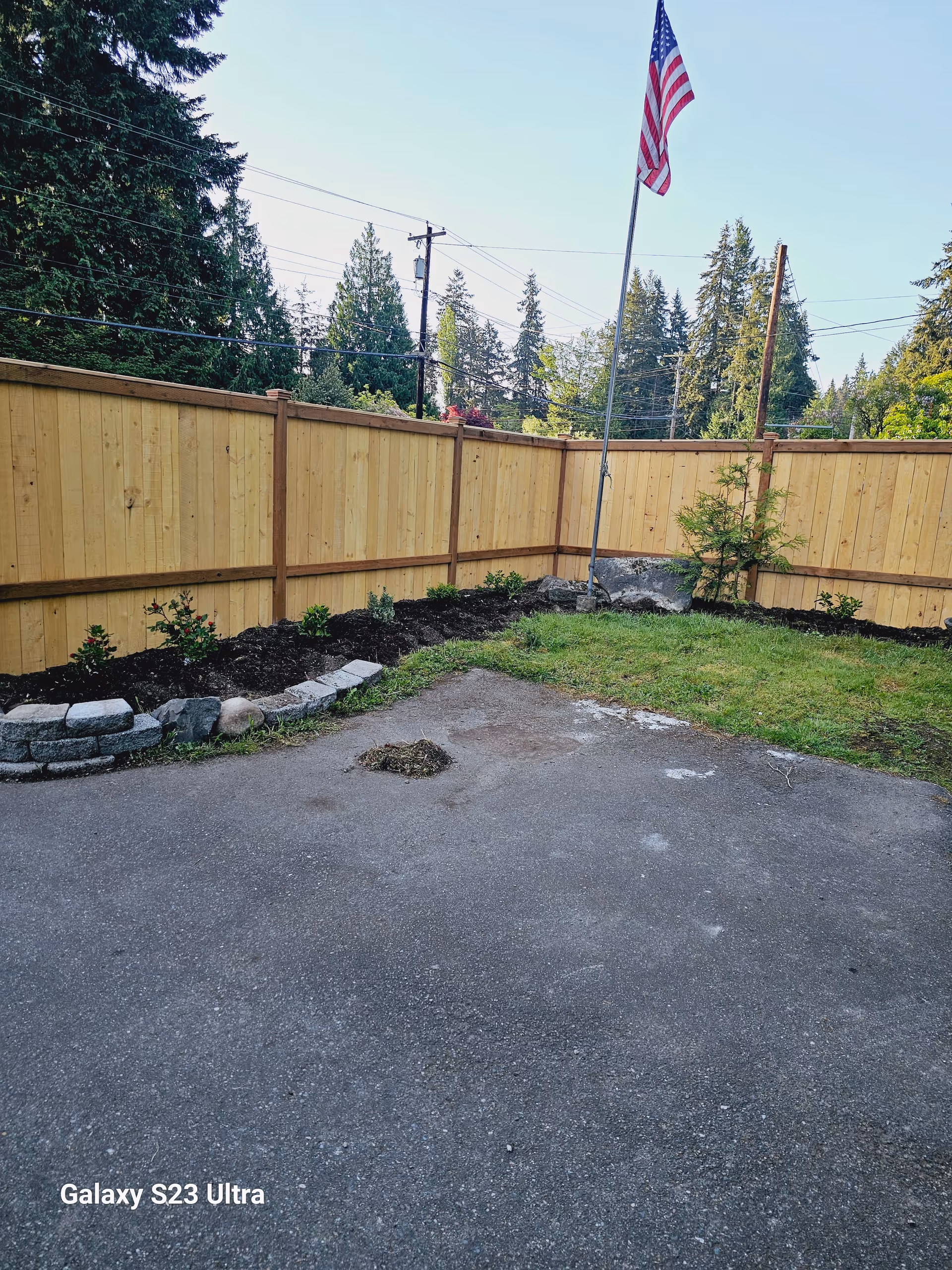 Empty backyard with wooden fence, flagpole, and landscaped border along the edge.