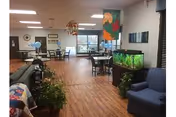 Spacious senior living lounge with seating, tables, plants, and a large fish tank.