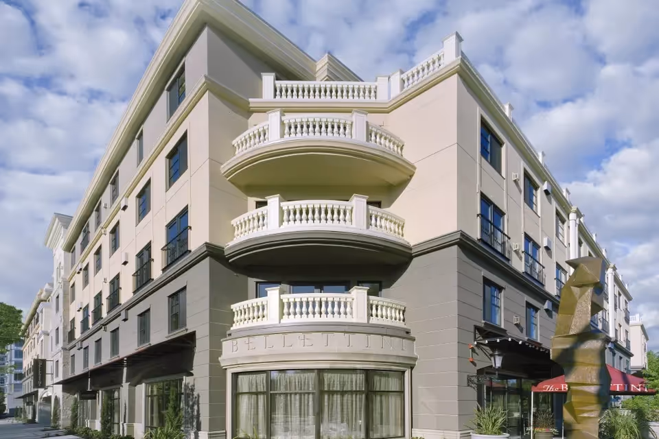 Exterior view of The Bellettini, a multi-story senior living facility with beige and gray walls, multiple balconies with white railings, large windows, and a sculpture in front under a partly cloudy sky.