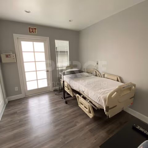 A hospital-style bed in a small gray patient bedroom beside an exit door.