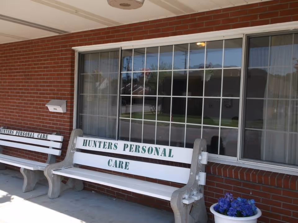 Two white benches with concrete legs placed outside a brick building under a covered porch. Both benches have the text 'HUNTERS PERSONAL CARE' painted in green. A large window with white grid frames is behind the benches, and a white flower pot with purple flowers is on the ground near one bench.
