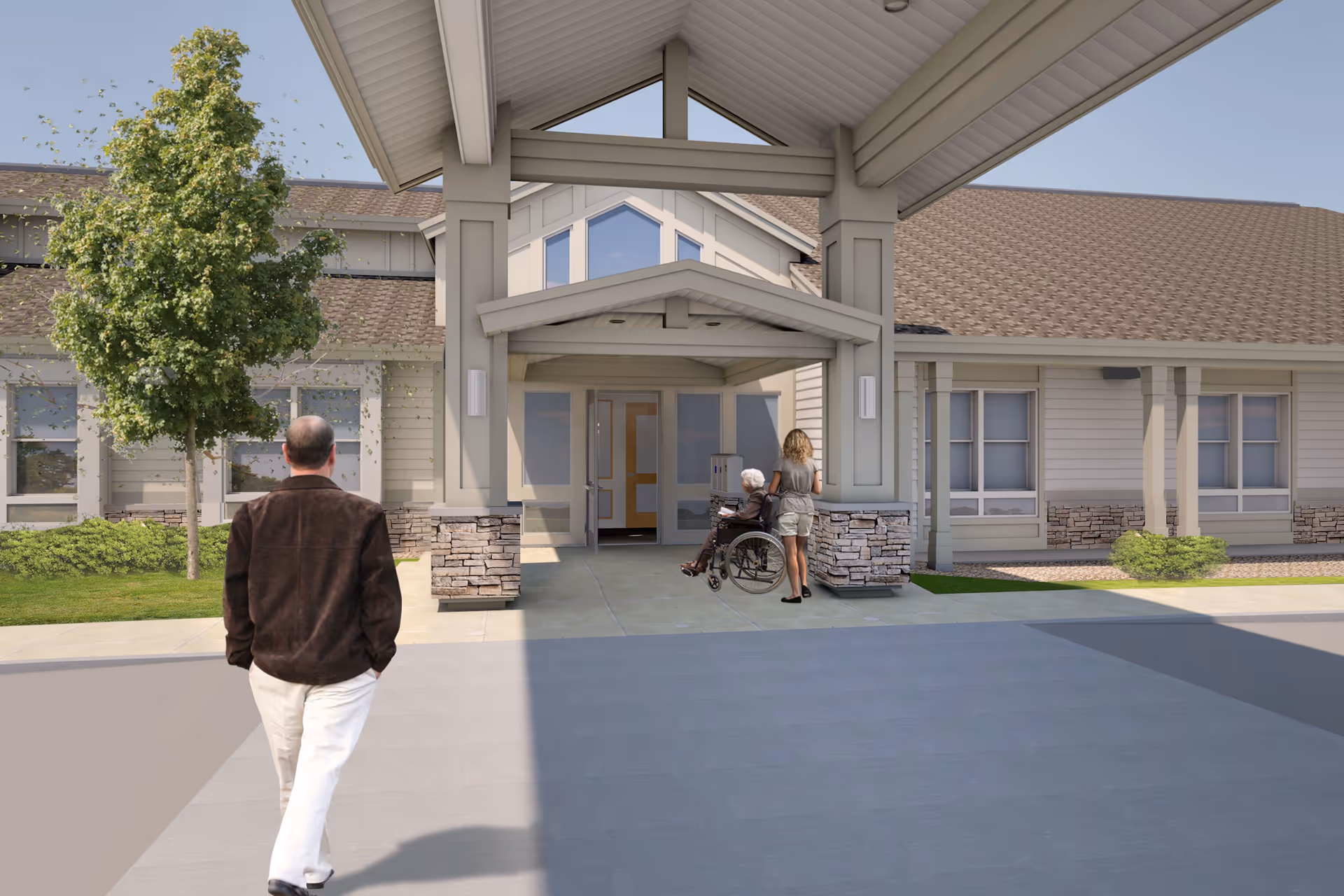 Entrance of a senior living facility with a covered drop-off area. A man is walking towards the entrance, and a woman is pushing an elderly person in a wheelchair towards the door. The building has beige siding with stone accents and a pitched roof. There is a tree and some greenery on the left side.