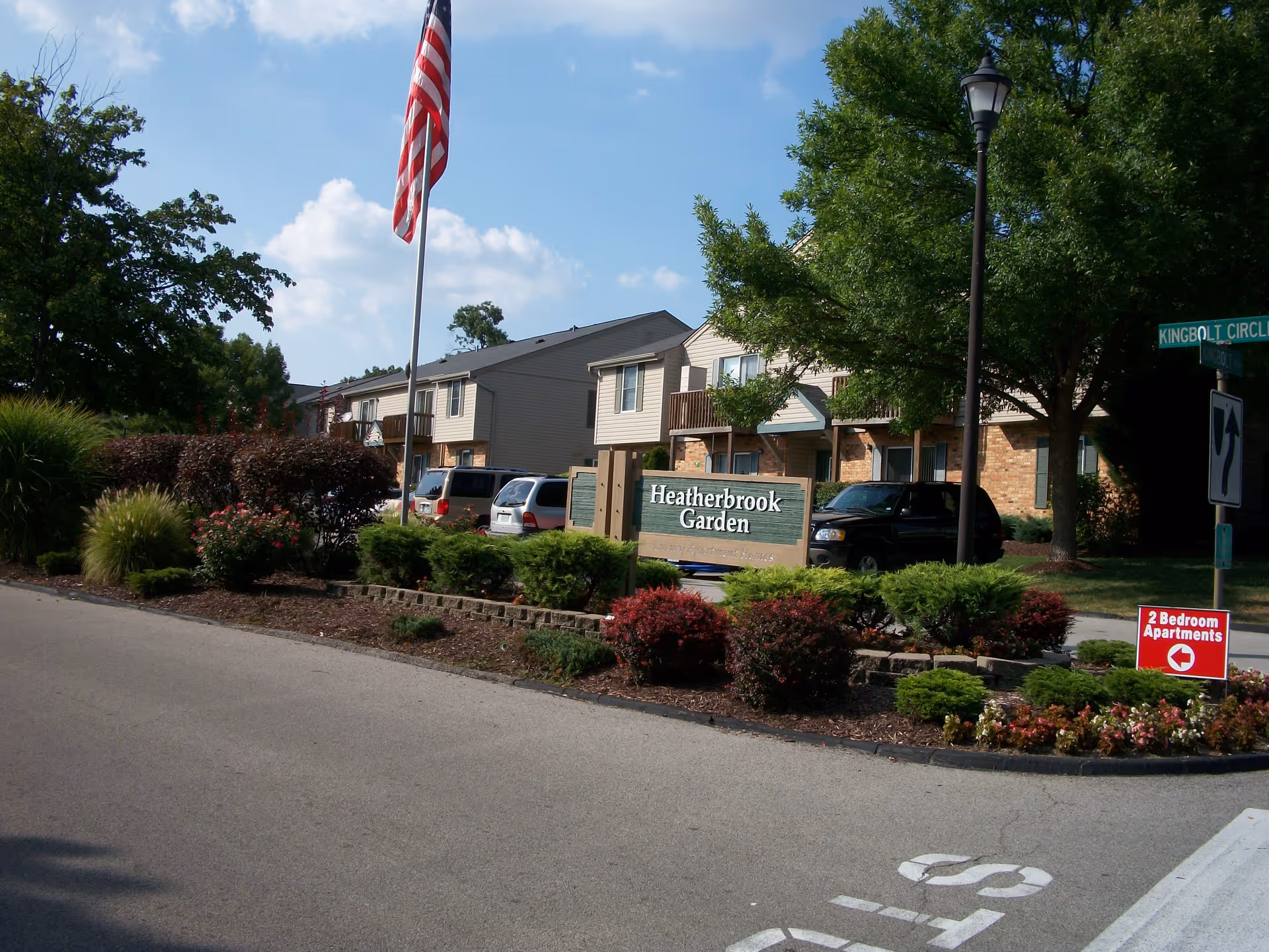 Exterior view of Heatherbrook Garden luxury apartment homes with a landscaped entrance, an American flag on a flagpole, several parked cars, and a street sign for Kingbolt Circle.