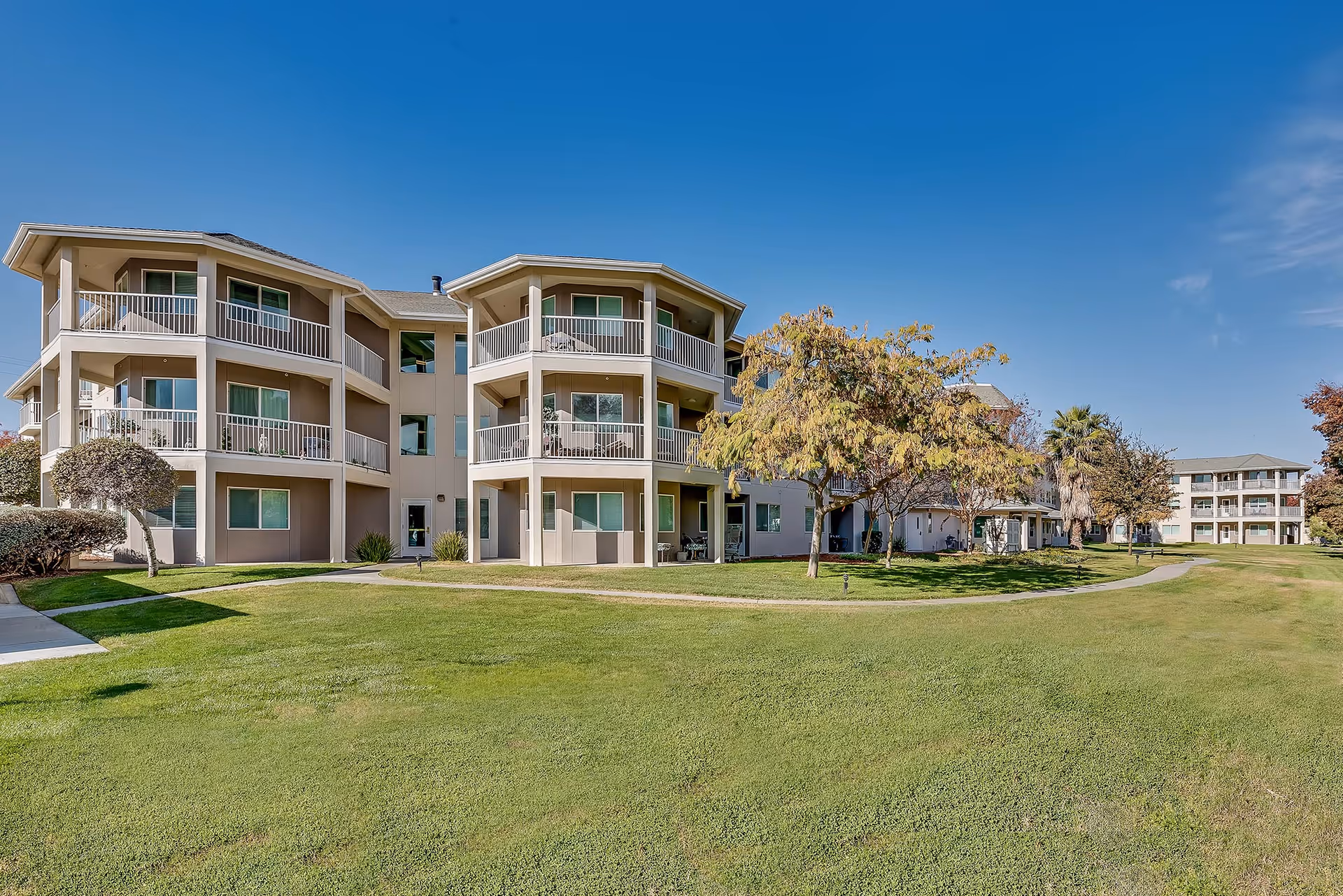 Three-story beige apartment-style senior living building with balconies overlooking a well-manicured lawn and walking paths.