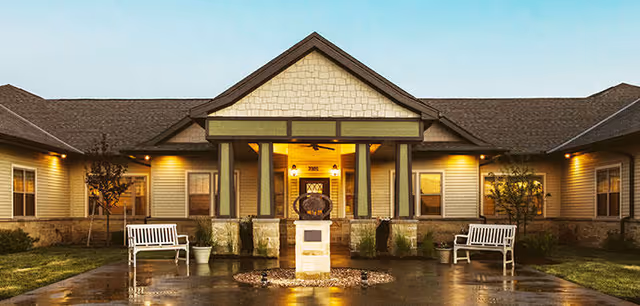 Exterior view of Silverado Lee's Summit Memory Care Community building at dusk with warm lights illuminating the entrance. The building has a peaked roof with stone and siding facade. There are two white benches and some small trees on either side of the entrance walkway.