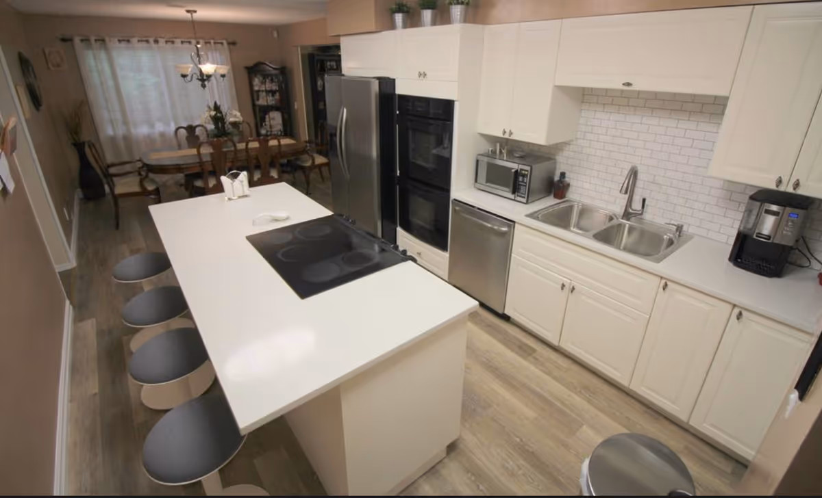 A modern kitchen with white cabinets, a white island countertop with an electric stove, and four gray stools. The kitchen features a double sink, a microwave, a dishwasher, a stainless steel refrigerator, and a coffee maker. In the background, there is a dining area with a wooden table and chairs, a chandelier, and a window with curtains.