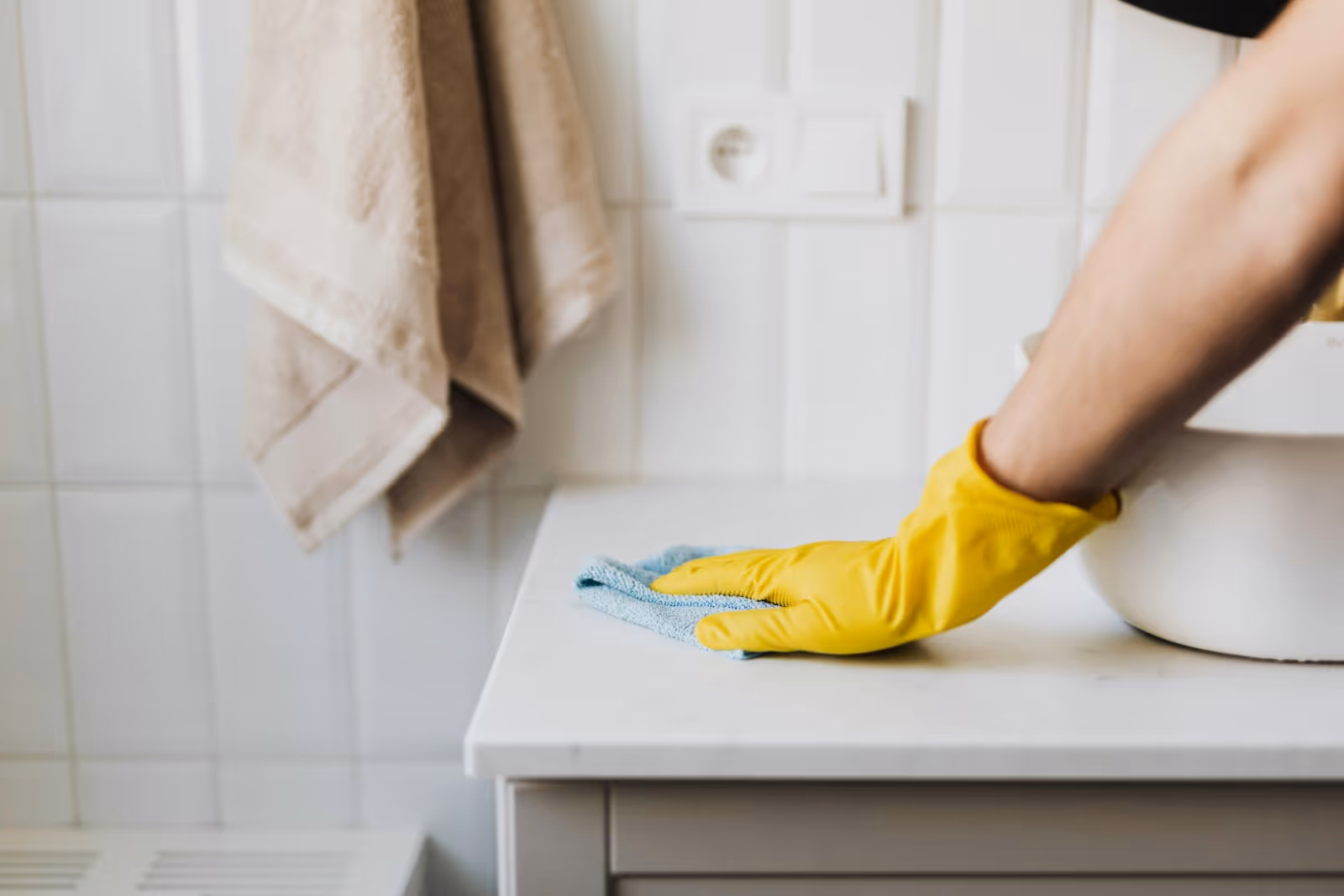 Gloved hand cleans a bathroom sink countertop with a blue cloth.