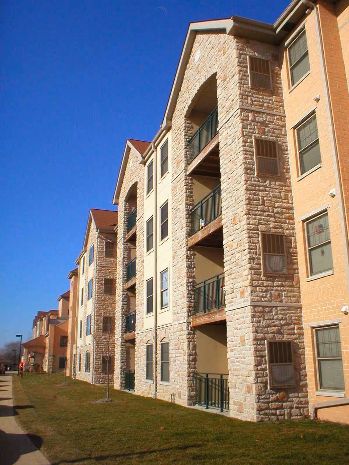 Stone-faced multi-story apartment building with balconies and a grassy lawn under a clear blue sky.