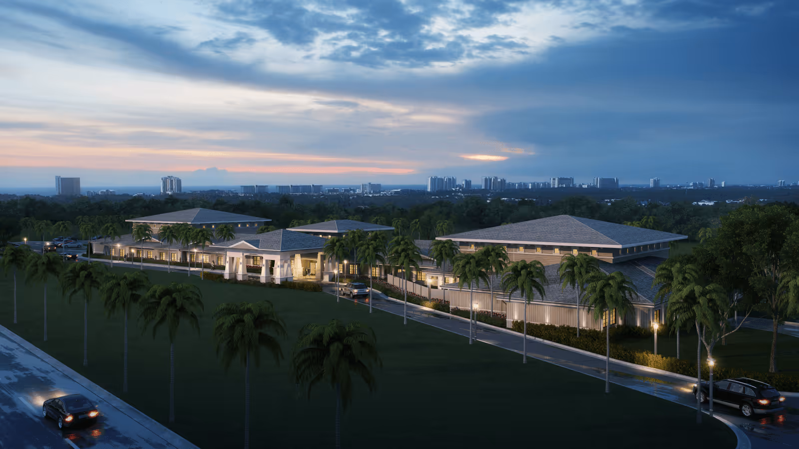 Aerial view of Seagrass Village of Panama City Beach senior living facility at dusk, showing multiple single-story buildings with pitched roofs surrounded by palm trees and greenery, with a city skyline in the background and cars parked along the driveway.