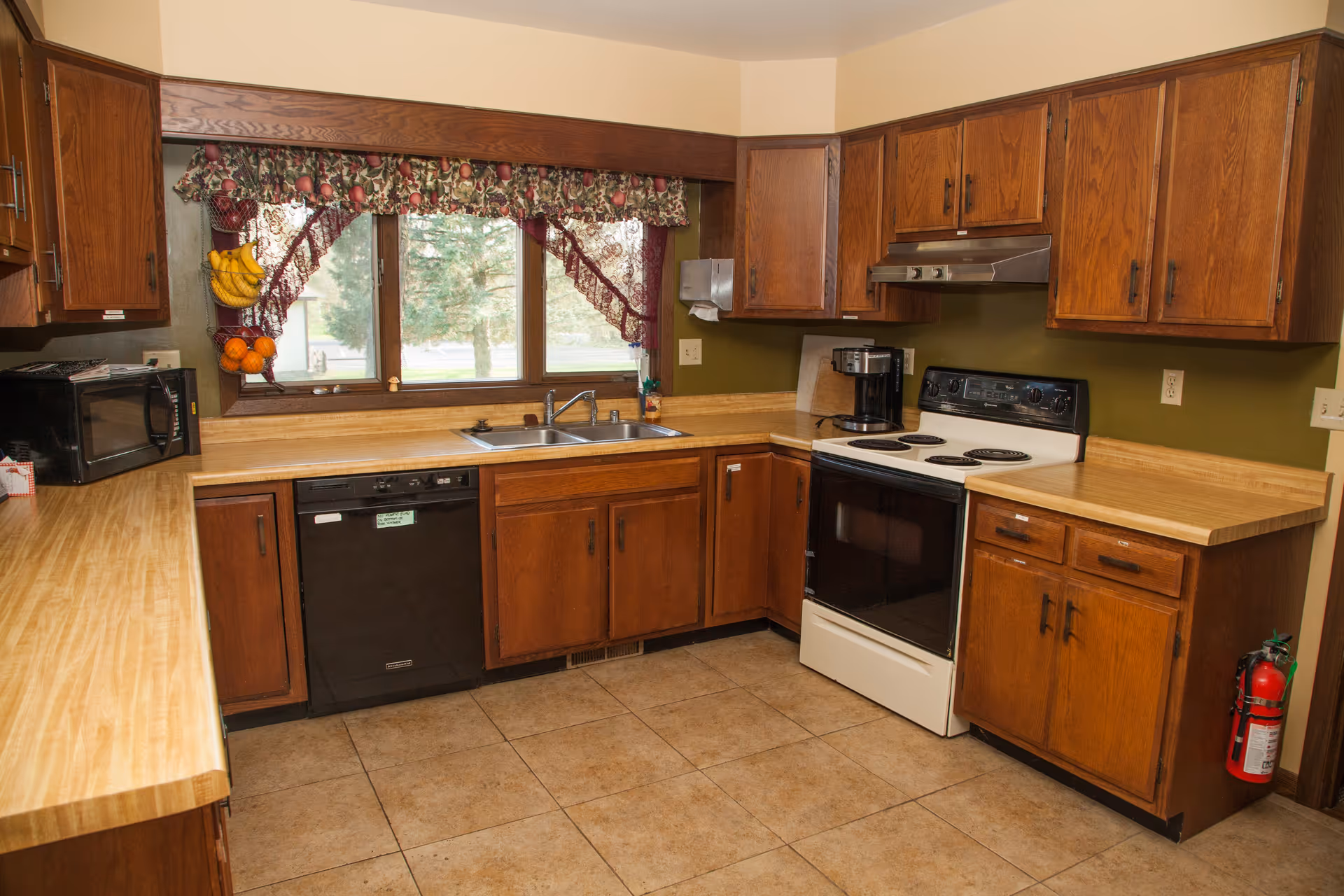 Wooden cabinets surround a kitchen with stove, sink, dishwasher, microwave, and window view.