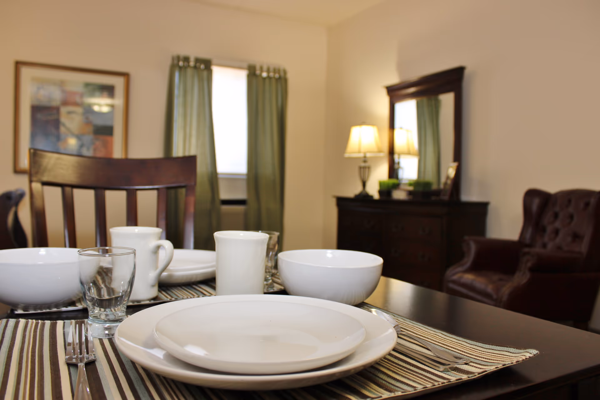 A dining table set with white plates, bowls, mugs, and a glass on striped placemats. In the background, there is a wooden chair, a window with green curtains, a framed picture on the wall, a dark wooden dresser with a mirror, a lamp, and a brown leather armchair.