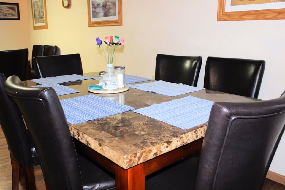 A dining table with a marble top surrounded by six black leather chairs. The table is set with blue striped placemats and a centerpiece featuring a small vase with colorful artificial flowers and salt and pepper shakers. The background shows framed pictures on a light-colored wall.