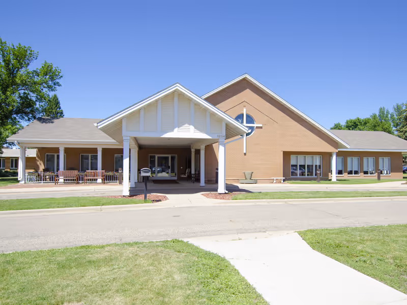 Front exterior view of a single-story brick building with a covered entrance supported by white columns. The building has multiple windows and a circular window with a cross design. There is a sidewalk and a road in front, with green grass and trees around the building under a clear blue sky.
