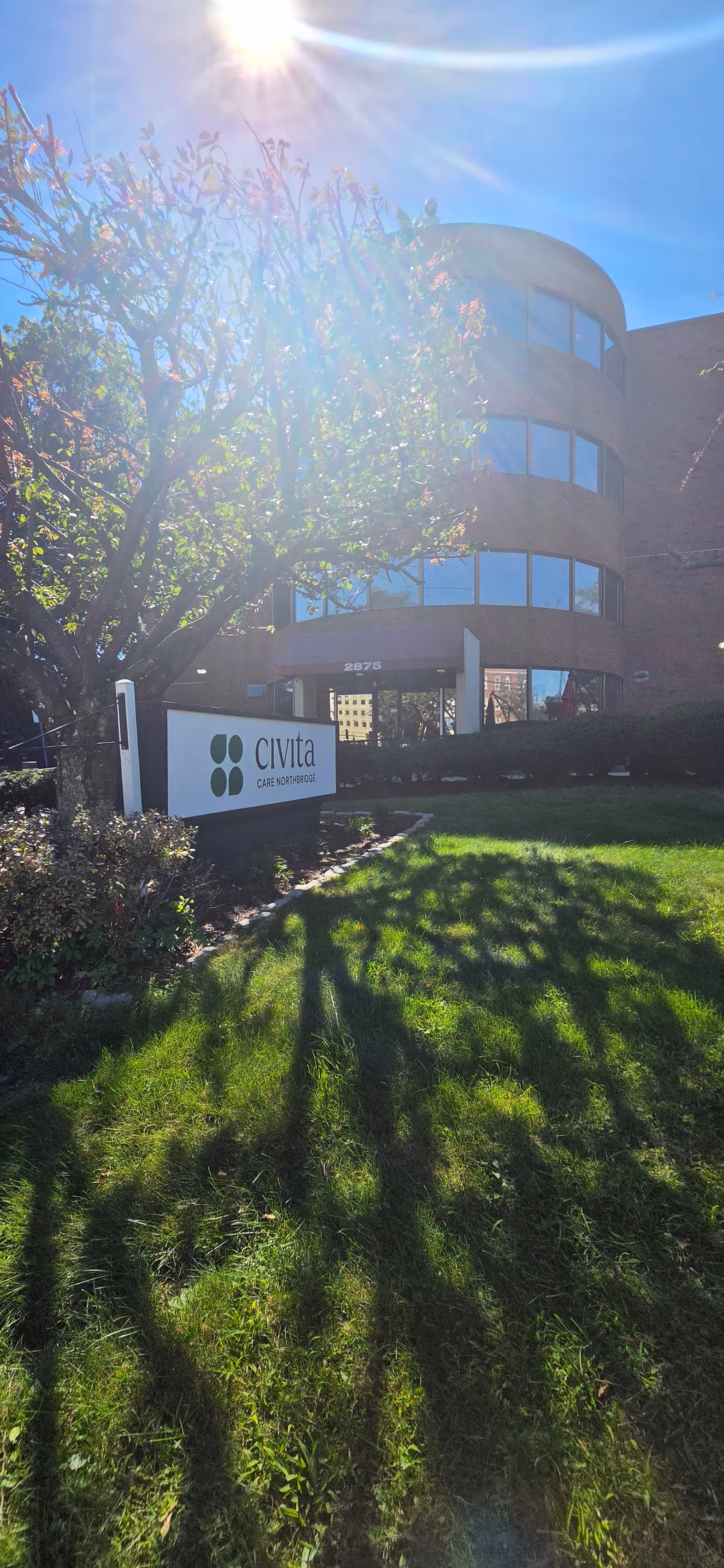 Exterior view of a curved glass-front building with a care facility sign and green lawn.