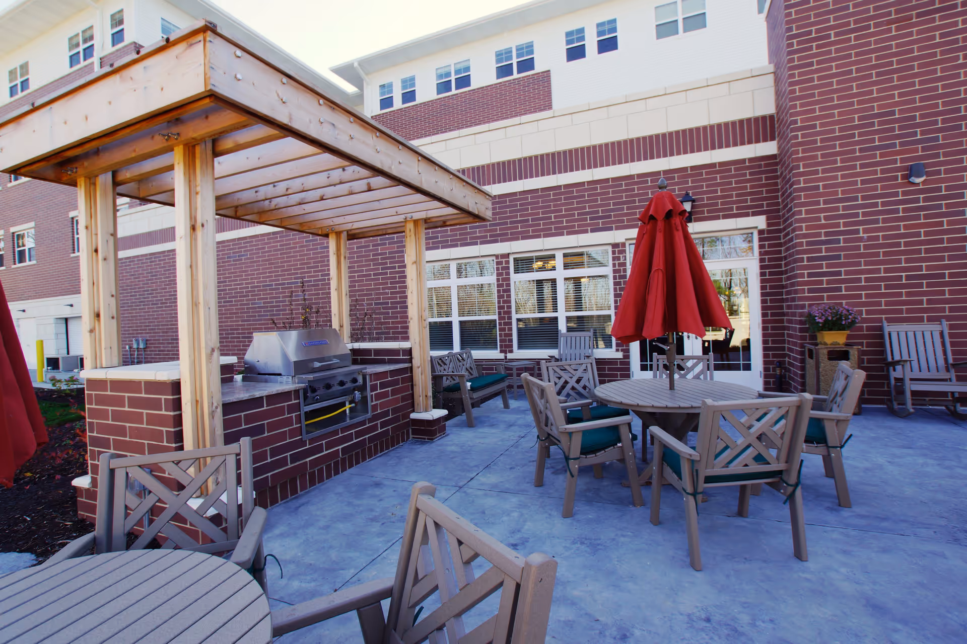 Outdoor patio area at Oak Hill facility featuring a built-in brick grill with a wooden pergola overhead, round tables with chairs, and a red umbrella. The background shows the brick exterior of the building with windows and doors.