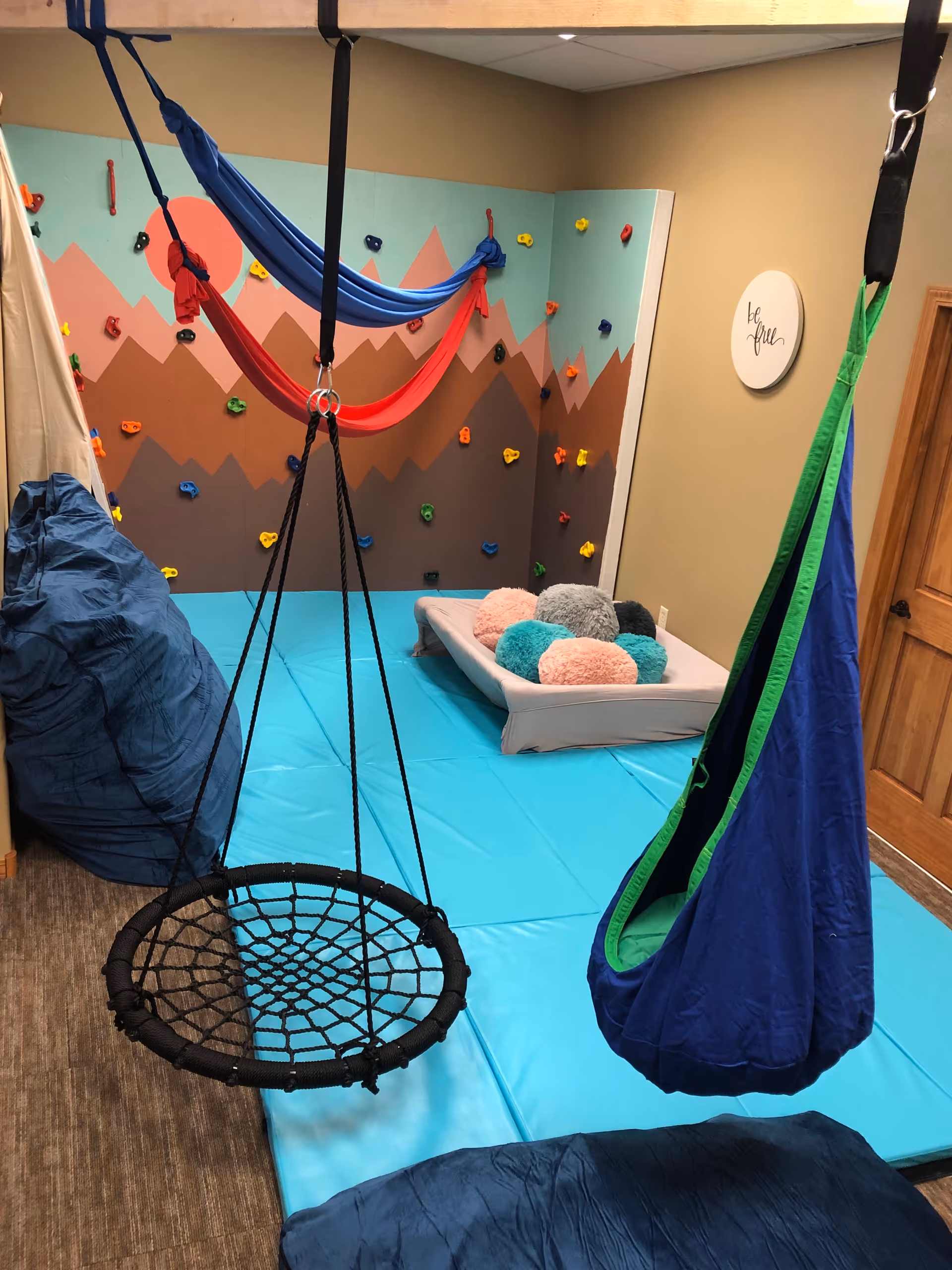 Indoor playroom with a blue padded floor, climbing wall, swings, and colorful cushions.