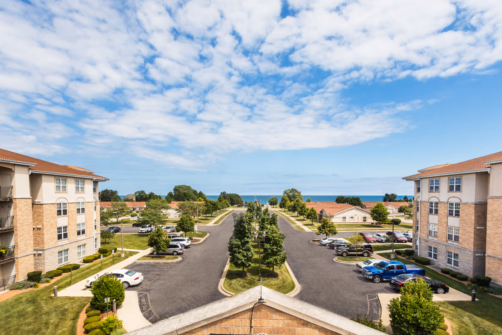 View of a senior living facility parking lot with two three-story buildings on either side, a tree-lined median in the center, and a clear blue sky with scattered clouds above. The ocean is visible in the distance beyond the buildings.