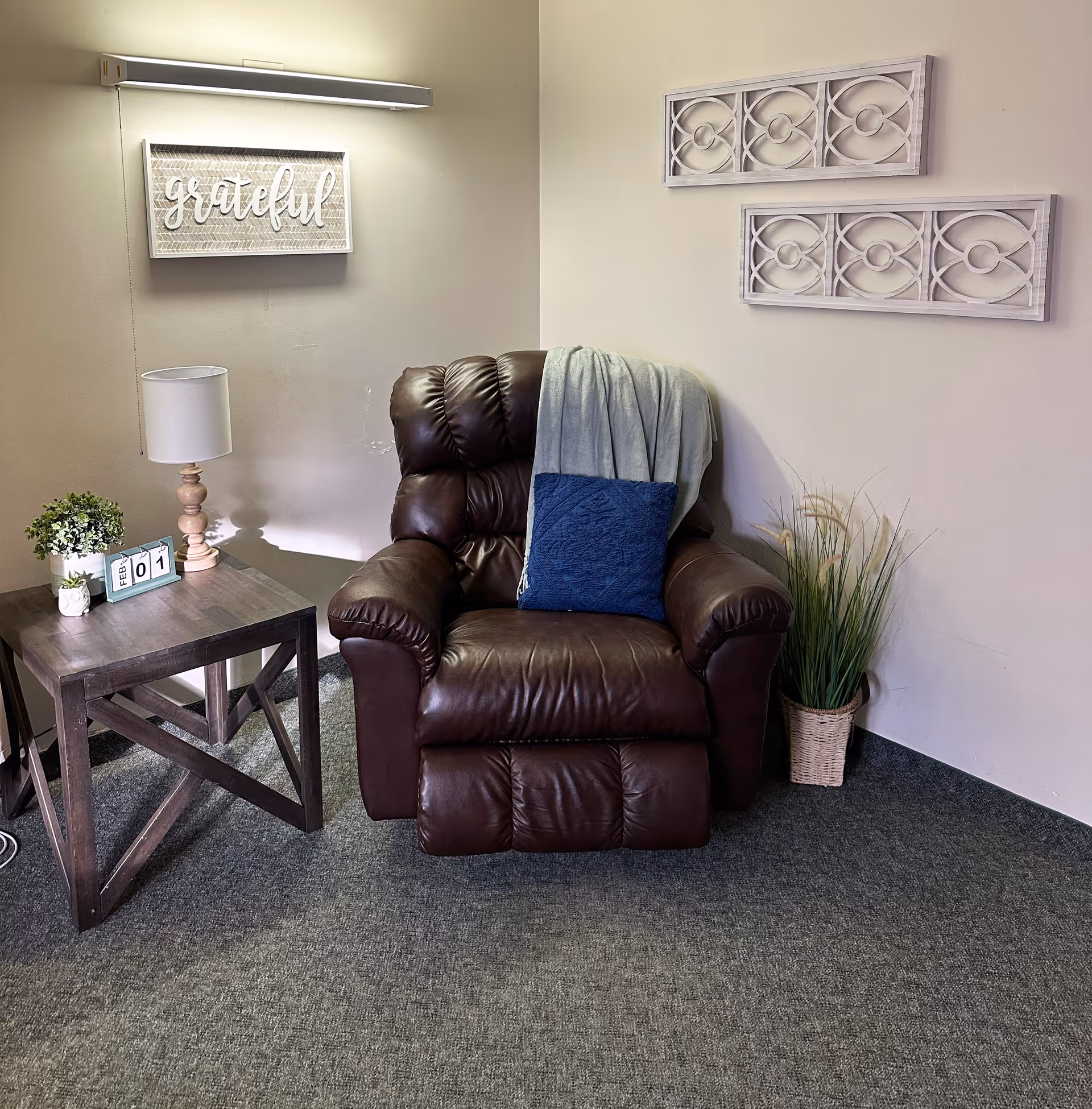 A cozy corner with a dark brown leather recliner chair draped with a light gray blanket and a blue cushion. Next to the chair is a wooden side table with a small lamp, a potted plant, and a calendar block showing February 1. On the wall above the chair is a decorative sign that says 'grateful' and two rectangular wall art pieces with circular patterns. A basket with tall grass-like plants is placed on the floor beside the chair.